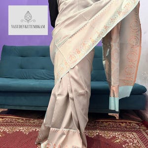 May include: A light beige saree with intricate floral and geometric patterns. The saree features a detailed border with orange and blue accents. The text "VASUDEVKUTUMBKAM" is visible on the left side of the image.