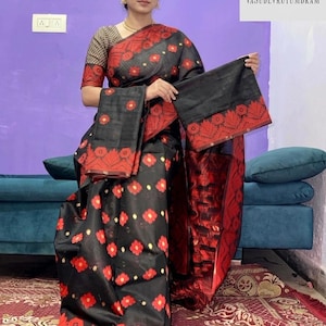 May include: A black saree featuring a repeating red floral pattern and small yellow dots. The garment includes a wide red border with intricate leaf motifs and a matching black blouse with a subtle geometric pattern on the sleeves and neckline.
