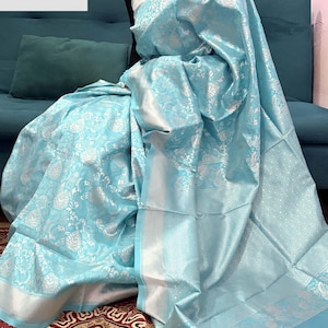 May include: A light blue sari with intricate silver floral patterns. The sari has a shimmering texture and a silver border. The text "VASUDEVKUTUMBKAM" is visible in the upper left corner. The sari is draped over a person sitting on a teal couch.