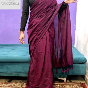 May include: A deep burgundy saree with a flowing drape. The saree is paired with a black long-sleeved top. The image includes the text "VASUDEVKUTUMBKAM" in a square frame. The saree has tassels at the end.