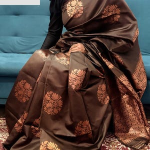 May include: A brown silk saree with a floral pattern in a lighter shade of brown. The saree has a decorative border and is draped over a person. The text "VASUDEVKUTUMBKAM" is visible in the upper left corner.