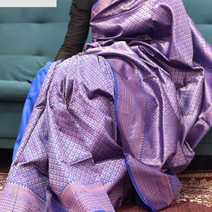May include: A draped purple and gold sari with intricate woven patterns. The sari features a detailed border with a blue trim. The fabric has a shimmering, textured appearance. The text "VASUDEVKUTUMBKAM" is visible in the upper left corner.
