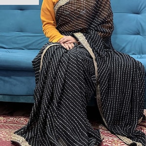 May include: Black and white striped saree with gold trim. The saree is draped over a person wearing a yellow long-sleeve shirt. The saree has a delicate pattern and is trimmed with gold along the edges. The text "VASUDEVKUTUMBKAM" is visible.