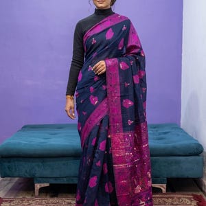 May include: A dark navy blue saree featuring a vibrant magenta floral pattern and a wide, ornate magenta border with intricate woven details. The garment is paired with a black long-sleeved mock neck top, creating a layered look with a distinct contrast.