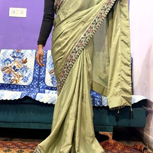 May include: A light green sari with intricate embroidered borders in red, green, and gold. The sari has a flowing drape and is embellished with delicate floral embroidery. The garment is worn over a black long-sleeved top.