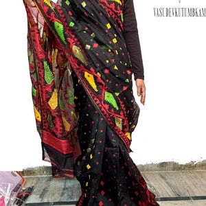 May include: Black sari with red, yellow, and green embroidered details. The sari features a repeating pattern of diamonds and floral designs. The text "VASUDEVKUTUMBKAM" is visible in the upper right corner.