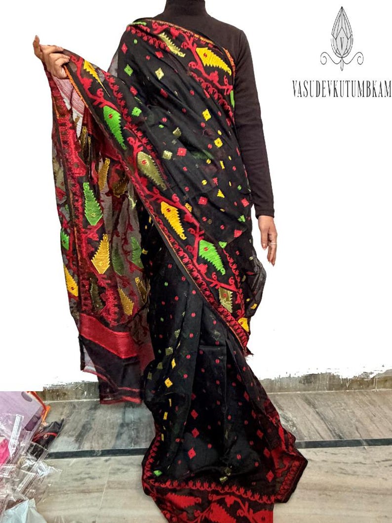 May include: Black sari with red, yellow, and green embroidered details. The sari features a repeating pattern of diamonds and floral designs. The text "VASUDEVKUTUMBKAM" is visible in the upper right corner.