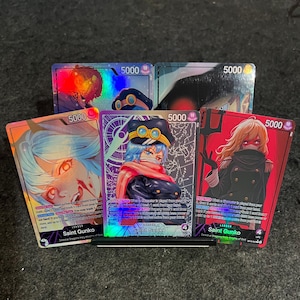 Pode incluir: Cinco cartas colecionáveis com ilustrações de personagens em estilo anime. Três cartas na fila da frente estão rotuladas como Saint Gunko com um nível de poder de 5000. As cartas exibem cores vibrantes, acabamentos holográficos e texto.