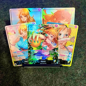 Puede incluir: Un conjunto de cinco cartas coleccionables con ilustraciones de estilo anime de un personaje llamado Nami. Las cartas tienen un brillo holográfico e incluyen el número "5000". La carta central muestra el nombre "Nami" y "Straw Hat Crew".