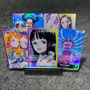 Puede incluir: Cinco cartas coleccionables de One Piece con Nico Robin, cada una con un nivel de poder de 5000. Las cartas muestran varias ilustraciones coloridas del personaje, incluyendo escenas con otras figuras, todas etiquetadas con el nombre Nico Robin y texto.