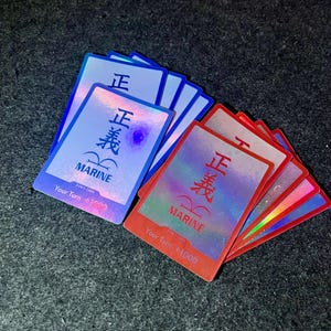 Puede incluir: Un conjunto de cartas coleccionables holográficas, algunas azules y otras rojas. Cada carta presenta caracteres japoneses, la palabra "MARINE" y el texto "Your Turn +1000". Las cartas tienen un acabado brillante e iridiscente.