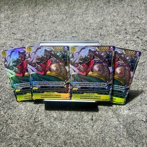 Peut inclure: Quatre cartes à collectionner Marshall D. Teach identiques du jeu One Piece, chacune avec un niveau de puissance de 10000, une bordure jaune et une illustration du personnage portant une chemise rouge, un pantalon vert et une barbe sombre.