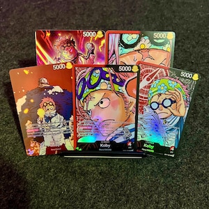 May include: A collection of five trading cards featuring colorful illustrations of a character named Koby. Each card has a 5000 value indicator. The cards are red, pink, and purple with various action poses.