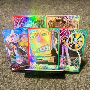 Op de afbeelding: Een verzameling holografische ruilkaarten met personages uit de anime-serie "One Piece". Elke kaart toont levendige illustraties, karakternamen en spelgerelateerde tekst. De kaarten hebben een waarde van "5000".