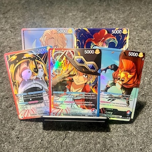 Puede incluir: Seis coloridas cartas coleccionables con ilustraciones de estilo anime. Cada carta muestra un personaje con diferentes fondos y texto, incluyendo el número "5000". Las cartas se exhiben en un soporte negro, mostrando su diseño.