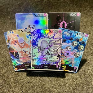 Puede incluir: Un conjunto de cartas coleccionables con diversos personajes y diseños. Las cartas muestran el número "5000" en las esquinas superiores. Las cartas tienen un efecto holográfico y presentan el nombre "Enel" y el texto "Sky Island".