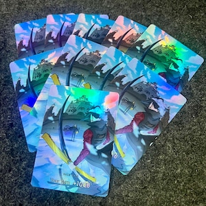 May include: A collection of holographic trading cards featuring a character in a red and purple outfit with a sword. The cards have a blue and white background with a castle and the text "Your Turn +1000".