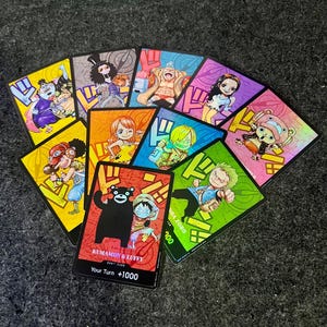 Könnte beinhalten: Eine Sammlung farbenfroher Sammelkarten mit verschiedenen Anime-Charakteren. Jede Karte hat ein einzigartiges Design und eine Charakterillustration. Eine Karte zeigt den Text "KUMAMON & LUFFY" und "Your Turn +1000". Die Karten sind fächerförmig angeordnet.