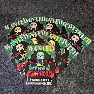 Puede incluir: Un conjunto de cartas coleccionables con una calavera coronada, el texto "WANTED DEAD OR ALIVE" y "THE SKBROOK". Las cartas tienen un efecto holográfico e incluyen el texto "Your turn +1000 MARINE".