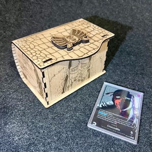 Puede incluir: Una caja de madera con tapa con bisagras, con un diseño cortado con láser y un emblema decorativo. La caja tiene un patrón texturizado e ilustraciones detalladas. Una carta coleccionable en una funda protectora está al lado.