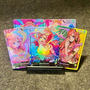 Könnte beinhalten: Eine Sammlung farbenfroher Sammelkarten mit Anime-Illustrationen der Figur Shirahoshi. Die Karten zeigen die Zahl 5000 und englischen Text. Die Karten werden auf einem schwarzen Ständer präsentiert.