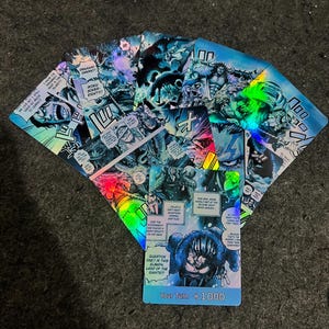Puede incluir: Una colección de tarjetas coleccionables rectangulares con ilustraciones holográficas estilo manga en tonos azules y negros. Las tarjetas muestran varios paneles de personajes, globos de texto con inglés y una tarjeta inferior etiquetada Your Turn +1000.