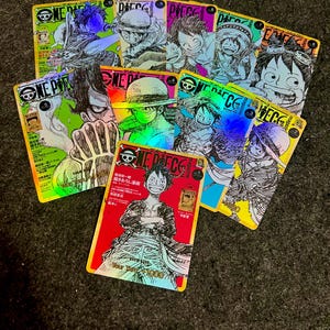 Puede incluir: Un conjunto de tarjetas coleccionables holográficas con ilustraciones de manga. Las tarjetas muestran varios personajes y el logotipo "ONE PIECE". Las tarjetas tienen un acabado brillante y colores vibrantes, incluyendo rojo, verde y azul.