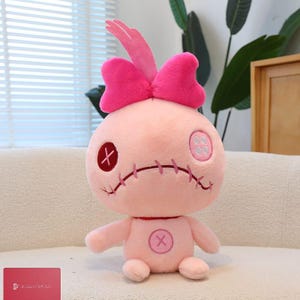Puede incluir: Una muñeca de peluche rosa con un gran lazo rosa en la cabeza. La muñeca tiene rasgos cosidos, incluyendo una cruz roja para un ojo y una boca cosida. Una "X" roja está en su pecho.