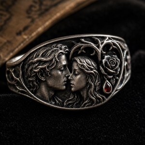 May include: A silver-toned metal ring featuring a detailed relief sculpture of two faces in profile facing each other. The design includes intricate vine-like branches, a sculpted rose, and a single small, teardrop-shaped red gemstone accent.