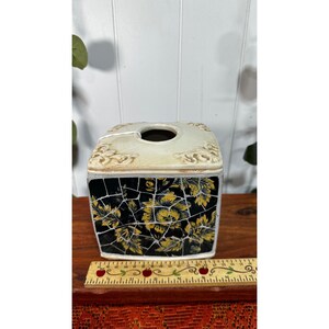 May include: A square tissue box cover featuring a black and yellow floral mosaic pattern on the sides and a cream-coloured top with decorative relief details. A wooden ruler is placed in front, showing a width of approximately 13 cm.