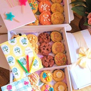 May include: Three stacked white cardboard boxes filled with assorted round biscuits, including chocolate chip, sprinkle-topped, and letter-shaped varieties. Each box includes a decorative greeting card and colourful star or stick candles.