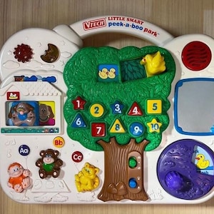 May include: A white VTech Little Smart Peek-a-boo Park plastic activity center featuring a large green tree with numbered shapes from 1 to 10, a house with a dog, a mirror, a purple spinning wheel with a duck, and various animal figures.