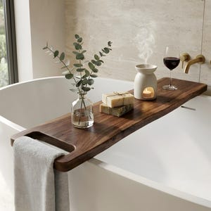 May include: A wooden bath tray with a live edge, holding a glass of red wine, a candle diffuser, and bars of soap. A vase with green sprigs and a gray towel complete the spa-like scene. The tray is set over a white bathtub.