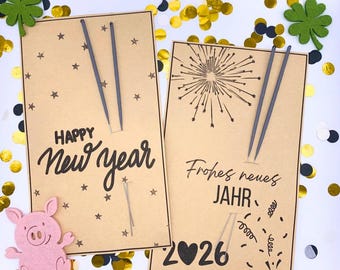 Könnte beinhalten: Zwei Neujahrskarten aus Kraftpapier mit Stern- und Feuerwerksillustrationen. Eine Karte sagt "Happy New Year" und die andere "Frohes neues Jahr" mit "2026". Dekorative Konfetti und rosa Schwein- und grüne Kleeblatt-Akzente.
