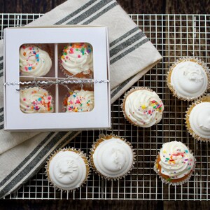 May include: A selection of cupcakes with white frosting and colorful sprinkles. Some cupcakes are in a white box, while others are on a metal cooling rack. A striped cloth is also present.