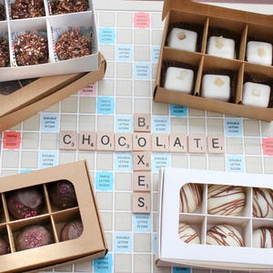 May include: Several boxes of gourmet chocolates are arranged on a Scrabble board spelling out "CHOCOLATE". The chocolates are in various shapes and colors, including dark chocolate truffles and white chocolate bonbons. The boxes are brown and white.