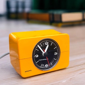Könnte beinhalten: Ein leuchtend gelber Krups Comfortime 3 Wecker im Retro-Stil. Die Uhr hat eine quadratische Form mit schwarzem Zifferblatt, weißen Zahlen und einem roten Sekundenzeiger. Die Uhr ist mit einem grauen Kabel angeschlossen.