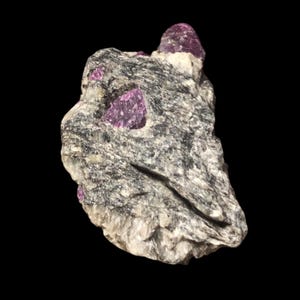 Könnte beinhalten: Ein rauer, grauer Felsformation mit eingebetteten, leuchtend rosa Kristallen. Der Felsen hat eine geschichtete Textur mit weißen und schwarzen Mineraleinschlüssen. Die Kristalle sind tiefrosa und kontrastieren mit dem grauen Stein.
