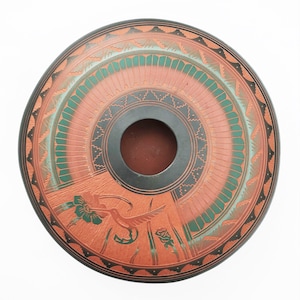 Op de afbeelding: Een ronde, handbeschilderde keramische kom met een centraal gat. De kom heeft een rood-oranje achtergrond met groene en zwarte geometrische patronen en een kolibrie- en bloemontwerp. De rand is zwart.