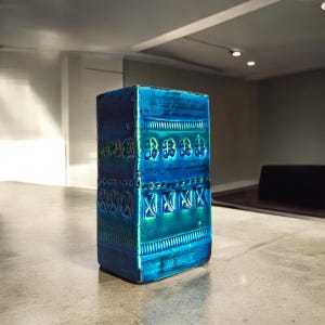 May include: A rectangular, turquoise and blue ceramic vase with embossed floral and geometric patterns. The vase has a glossy finish and is displayed on a neutral surface. The design features a mix of dark and light blue hues.