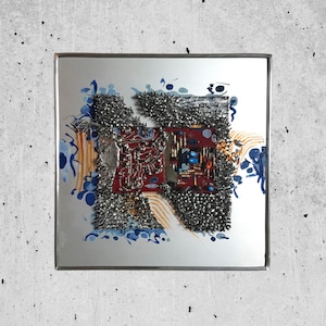 May include: Square artwork with a silver frame. The artwork features a central red and black design, surrounded by silver beads and accents of blue, orange, and white. The background is a light gray concrete wall.