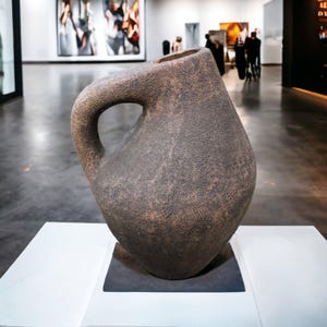 Könnte beinhalten: Eine große, strukturierte, braune Keramikvase mit Henkel. Die Vase hat eine einzigartige, asymmetrische Form und wird in einer Kunstgalerie auf einem weißen Sockel ausgestellt. Die Vase ist eine Skulptur.