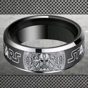 May include: Black ring with a silver band featuring the words "STAR WARS" in white. The center of the ring has a detailed illustration of Darth Vader's helmet. A stylish accessory for fans.