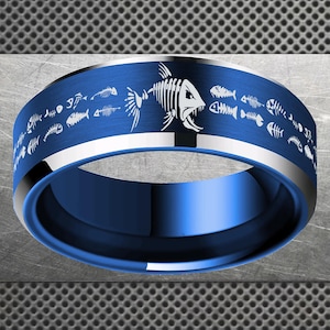 Puede incluir: Un anillo azul con un diseño de esqueleto de pez. El anillo tiene una banda azul cepillada con un borde plateado y un interior azul brillante. El diseño de esqueleto de pez blanco se repite alrededor del anillo.