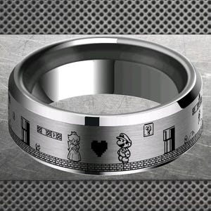 Puede incluir: Un anillo plateado con un acabado pulido. El anillo presenta un diseño grabado en negro de una escena de videojuego clásico, incluyendo personajes, ladrillos y puertas. El diseño del anillo es un homenaje a un videojuego popular.