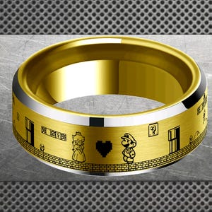 Puede incluir: Anillo dorado con una banda plateada y diseño grabado en negro. El anillo presenta un patrón de ladrillos, puertas y personajes de videojuegos. El anillo tiene un acabado pulido y una banda ancha.