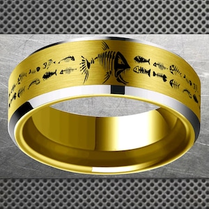 Puede incluir: Un anillo dorado con acabado cepillado, con un patrón repetido de esqueletos de peces negros. El anillo tiene una banda plateada en los bordes superior e inferior. El anillo es una toma de primer plano.