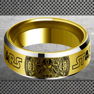 May include: Gold-toned ring featuring a black Darth Vader design and the words "WARS" and "STAR" in black. The ring has a silver band and a polished gold interior. A stylish accessory for fans of the iconic space saga.