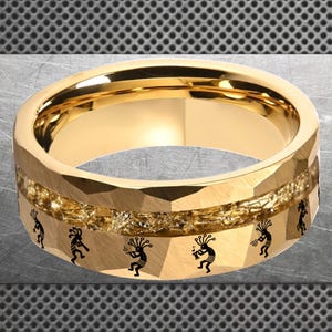 Puede incluir: Un anillo dorado con un exterior facetado y una banda central de material texturizado. La banda presenta figuras negras de Kokopelli. El anillo tiene un interior pulido y un diseño geométrico.
