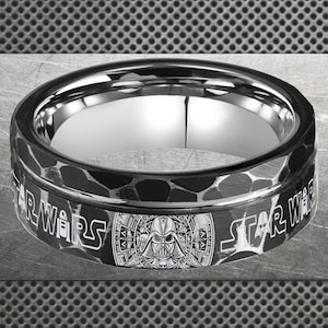 May include: A black and silver ring featuring the Star Wars logo and Darth Vader's helmet. The ring has a textured band with the words "STAR WARS" in white lettering. The inner band is polished silver.